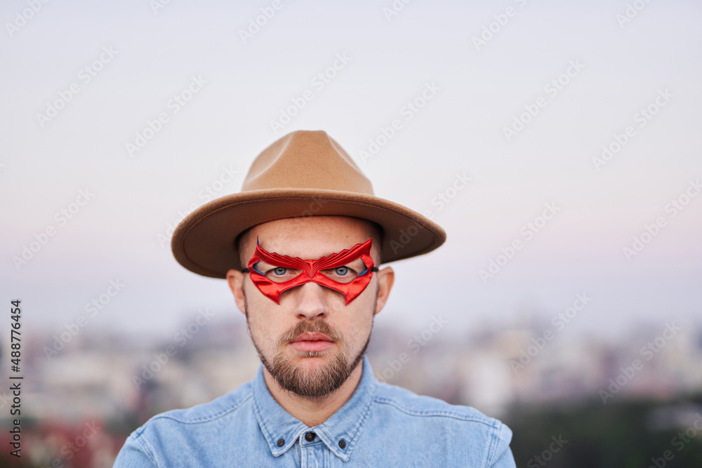 Man power, justice or superpower concept. Closeup portrait of bearded serious male in red superhero eyemask, brown hat and jeans shirt posing in front of the camera with city view. High quality image