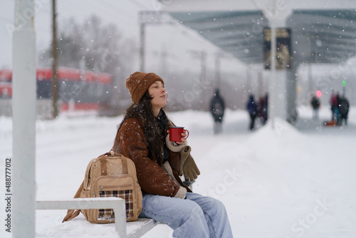 Tired but satisfied Asian traveler woman sitting on bench on snow-covered platform with mug of hot fragrant tea enjoying winter snowy weather while waiting for arrival of train. Selective focus