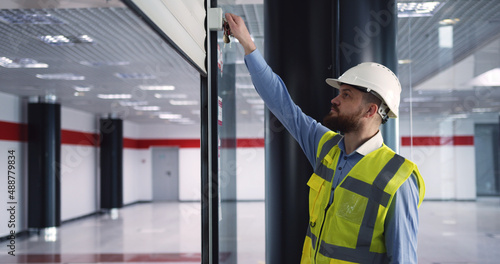 Male chief engineer in hardhat and vest standing inside commercial building construction site, closing an automatic roller door before leaving work.