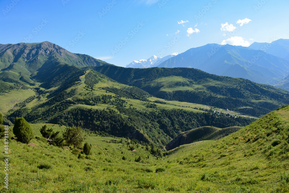 landscape of green mountains in spring with snowy peaks with blue sky and clouds and houses in the distance in spring in central asia kyrgyzstan