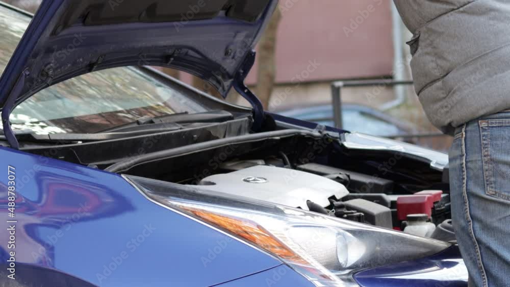 Car repairs and maintenance. A man in grey jacket is checking the oil level, outside, in the front of the garage. blue modern hybrid car. 4k video Car repairs and maintenance. A man in grey jacket is checking the oil level, outside, in the front of the garage. blue modern hybrid car. 4k video