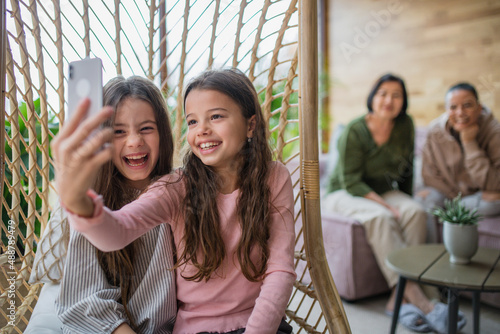 Happy little sisters sitting in wicker rattan hang chair indoors in conservatory and taking selfie. © Halfpoint