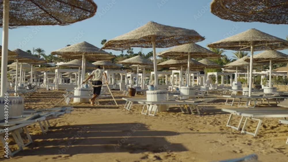 Beach staff walking among tents and lounges and flattening sand using special device. People working on resort. Summer holidays, vacation by the sea Beach staff walking among tents and lounges and flattening sand using special device. People working on resort. Summer holidays, vacation by the sea