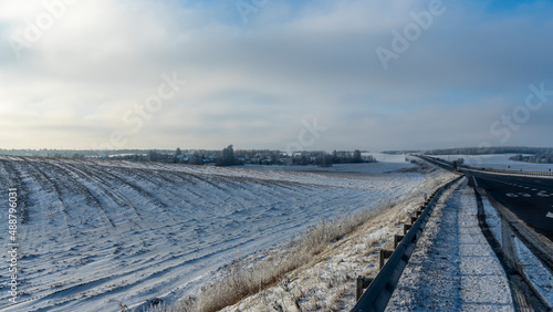 Winter road, agriculture field with snow in the countryside. Hoarfrost forest on a horizon. Winter landscape.