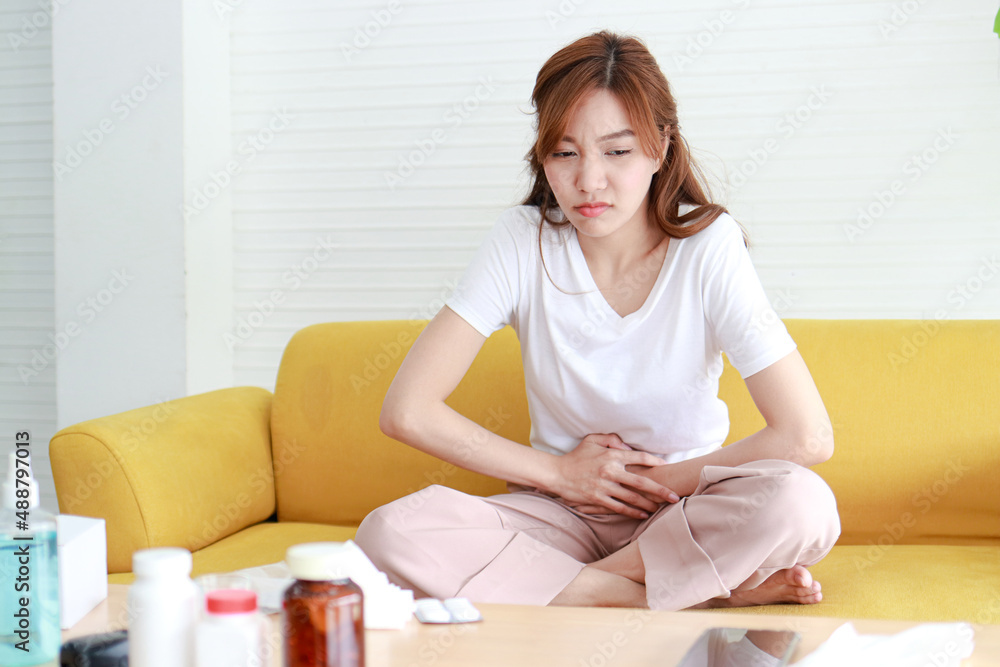 Beautiful Asian woman with lower abdominal pain. She sits on the sofa at home. Concept of treatment of infection, inflammation, digestive system, reproductive system in women. health care Beautiful Asian woman with lower abdominal pain. She sits on the sofa at home. Concept of treatment of infection, inflammation, digestive system, reproductive system in women. health care