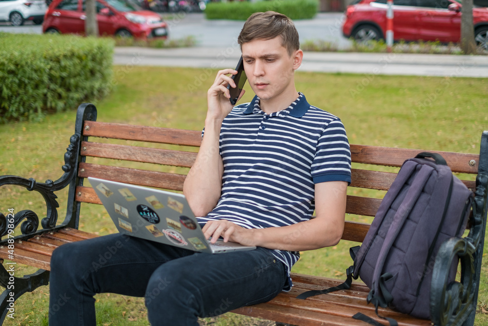 Fototapeta premium Young caucasian man sitting on the bench, working on the laptop and making
