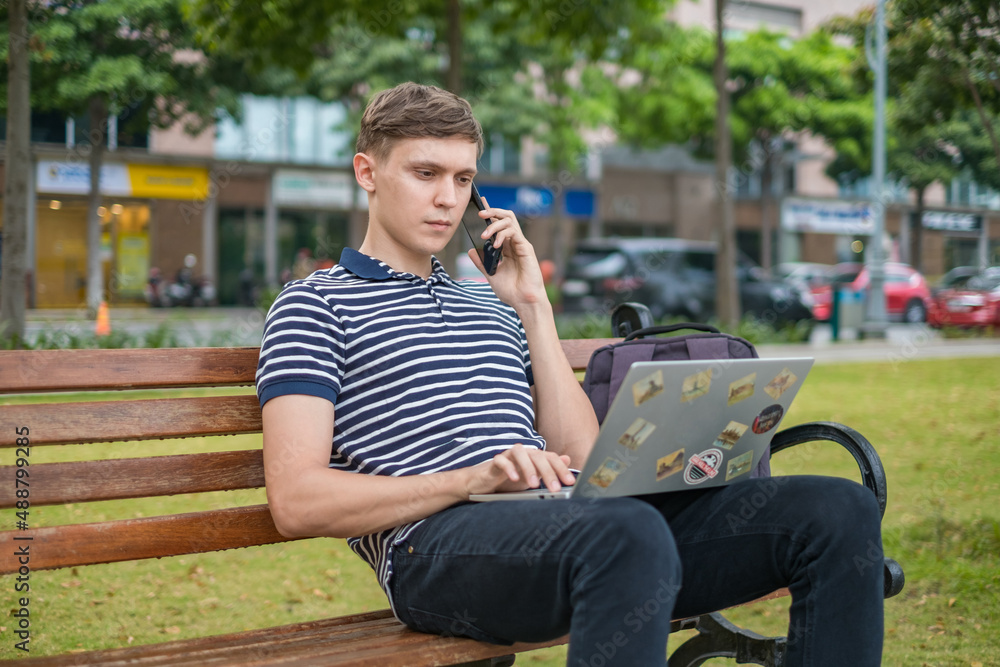 Young caucasian man sitting on the bench, working on the laptop and making Young caucasian man sitting on the bench, working on the laptop and making