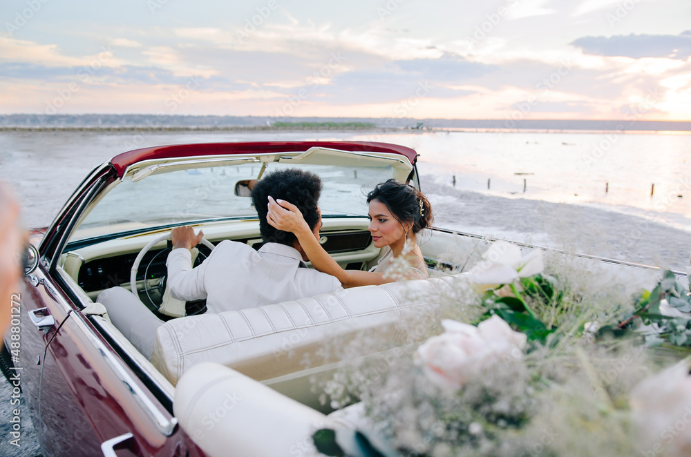 Beautiful couple at sunset on a summer wedding day. Romantic couple in love near their convertible. The groom is in a light suit, the bride is in a beautiful stylish dress.