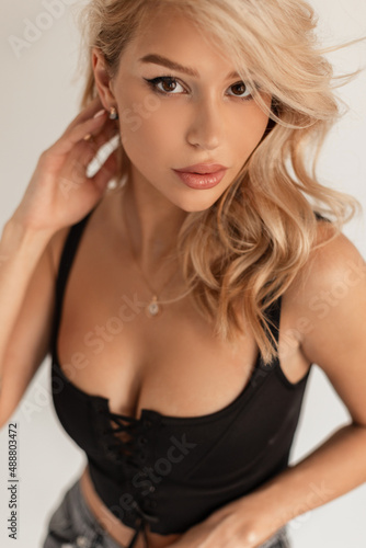 Glamorous portrait of a beautiful young girl with blond hair in a fashionable black sexy bra top in the studio on a white background looking at the camera