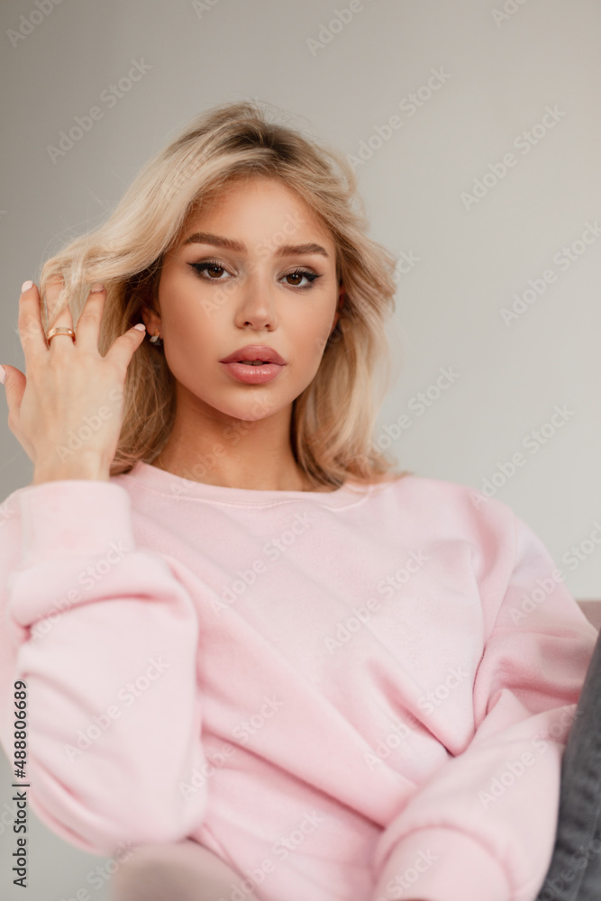 Stylish pretty young woman model with a cute face in a fashionable pink pullover with jeans sits in a pink armchair and straightens her blond hair. Girl looks at the camera Stylish pretty young woman model with a cute face in a fashionable pink pullover with jeans sits in a pink armchair and straightens her blond hair. Girl looks at the camera