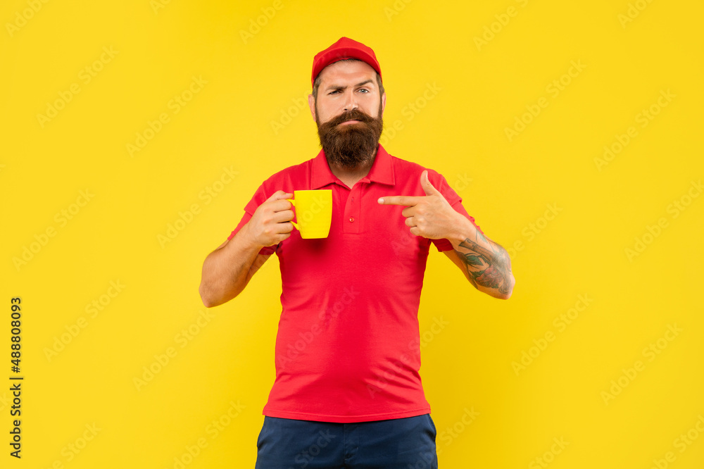 Serious frown bearded man in casual red cap and tshirt pointing finger at mug, coffee