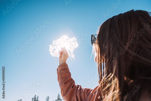A girl in black glasses holds a piece of ice in her hands and looks through it. Clear sunny weather, blue sky. © Margarita Timofeeva
