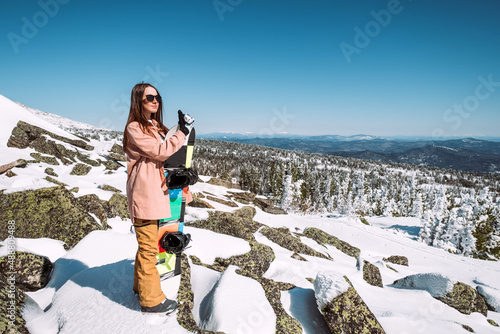 Russia. Sheregesh. Girl snowboarder in black sun goggles in winter in sunny weather outdoors among the Christmas tree and snow. The girl is standing with a snowboard in her hands and posing. 