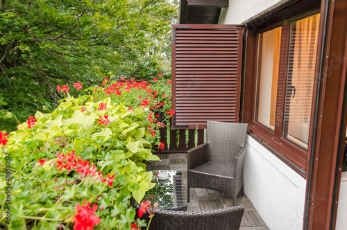 Outdoor Patio Furniture Set with Chairs and Table on the Balcony on a Sunny Day. Blooming Garden with Red Flowers Surround the Place. © Nikolaos