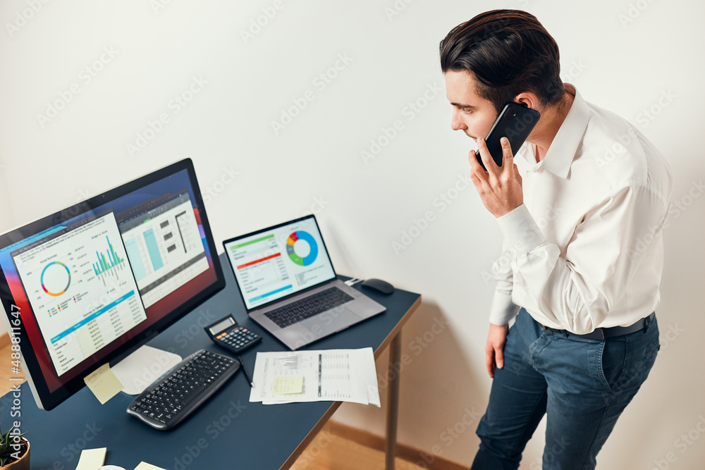 Man entrepreneur having business conversation on mobile phone. Businessman holding documents and smartphone working with data on charts and graphs in office. Man standing at desk Man entrepreneur having business conversation on mobile phone. Businessman holding documents and smartphone working with data on charts and graphs in office. Man standing at desk