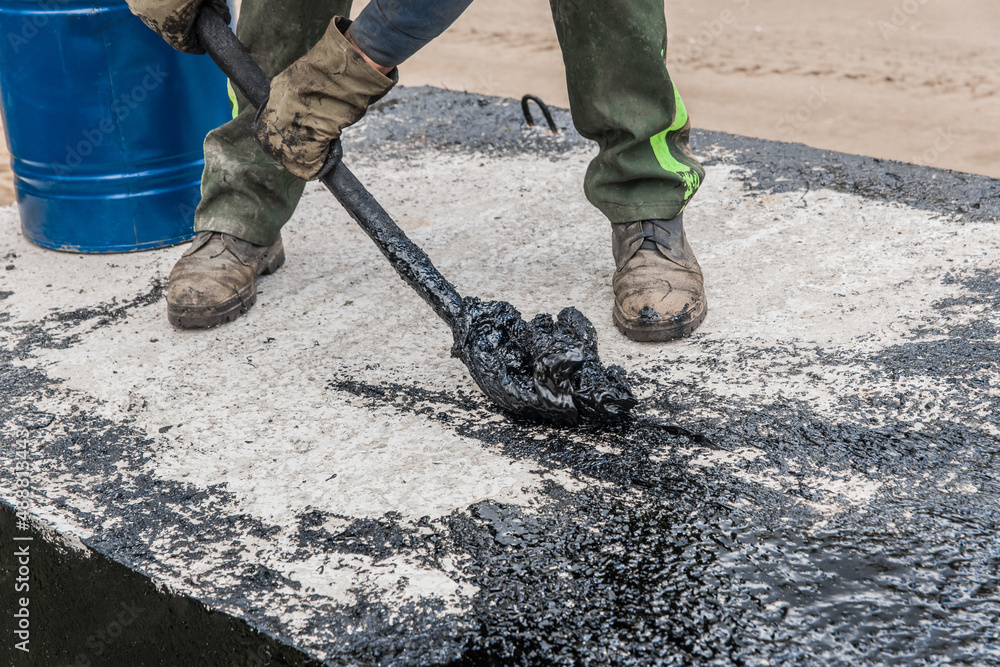 A construction worker is engaged in dirty manual work to repair and apply black bitumen waterproofing tar to the concrete surface of the structure A construction worker is engaged in dirty manual work to repair and apply black bitumen waterproofing tar to the concrete surface of the structure