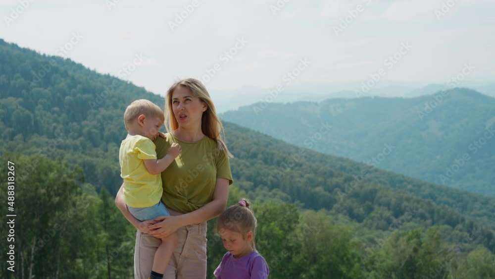 Pensive tired woman tourist with little unhappy children son and daughter stands against large green hills on sunny summer day slow motion Pensive tired woman tourist with little unhappy children son and daughter stands against large green hills on sunny summer day slow motion