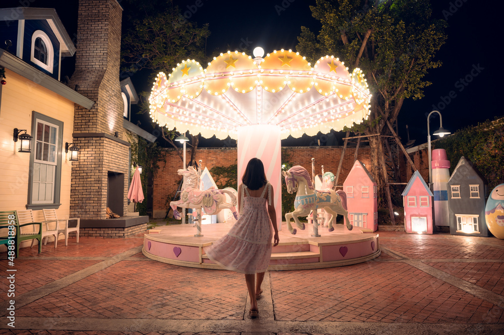 Back view of asian woman in dress walking to illuminated carousel at amusement park