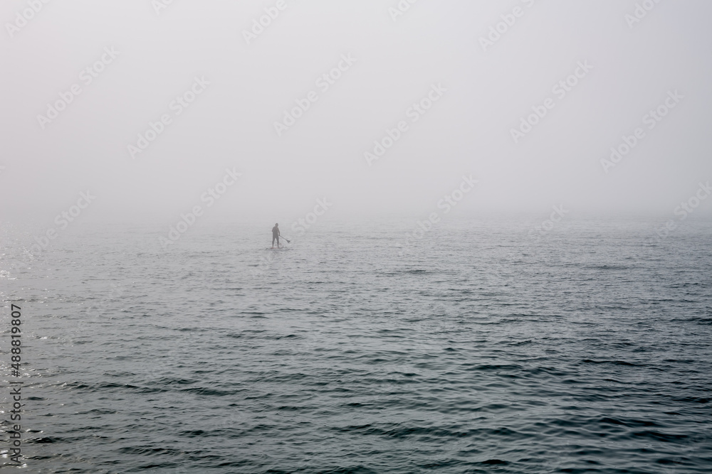 A man on a standing surf rows a paddle far out into the sea in the fog. A man on a standing surf rows a paddle far out into the sea in the fog.