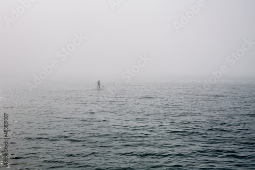 A man on a standing surf rows a paddle far out into the sea in the fog.