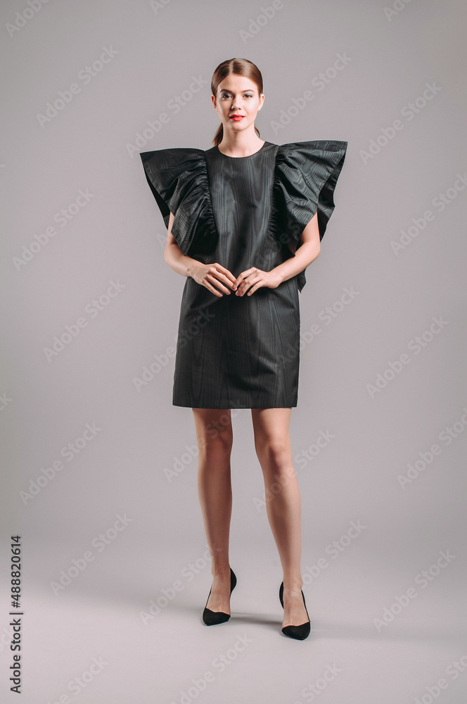 Fototapeta premium Oversize dress in black. Elegant cocktail midi dress with ruffles on massive shoulders. Beautiful young lady posing in studio. Elegant female fashion, summer evening look.