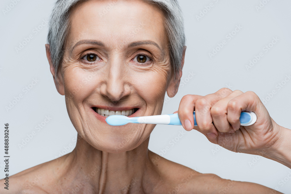 happy middle aged woman holding toothbrush and brushing teeth isolated on grey happy middle aged woman holding toothbrush and brushing teeth isolated on grey