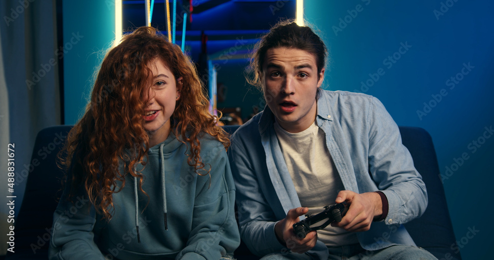 Funny young coupe playing video games. Woman gamer losing car racing competition, frowning and looking displeased. Two joyful people spending time together.