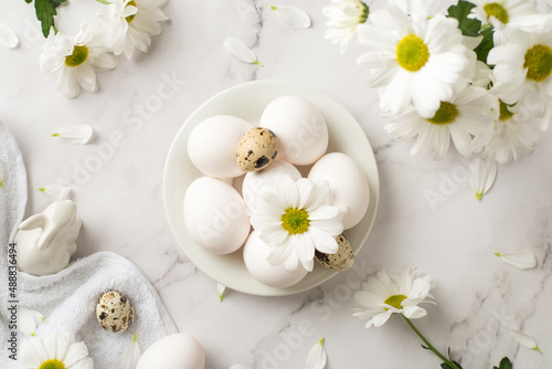 Top view photo of easter decorations festive table bouquet of chrysanthemum flowers ceramic easter bunny statuette towel and plate with white and quail eggs on isolated white marble texture background