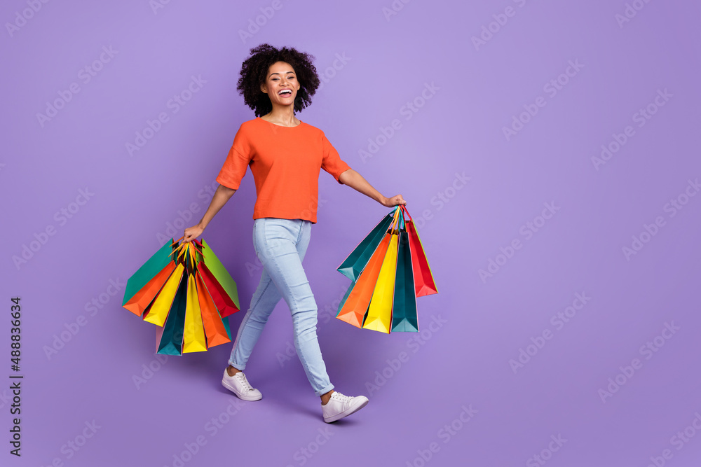 Full size photo of cute young brunette lady go with bags wear t-shirt jeans footwear isolated on violet background Full size photo of cute young brunette lady go with bags wear t-shirt jeans footwear isolated on violet background
