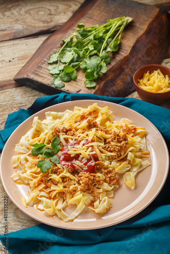 Pasta bolognese garnished with greens and cheese in a plate on a blue napkin on a wooden table next to a fork and greens on a board and grated cheese.