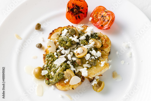 Cauliflower steak with spices, chimichurri sauce, almond flakes, olives, fried cherry tomatoes and capers on a white plate. Vegetarian food.