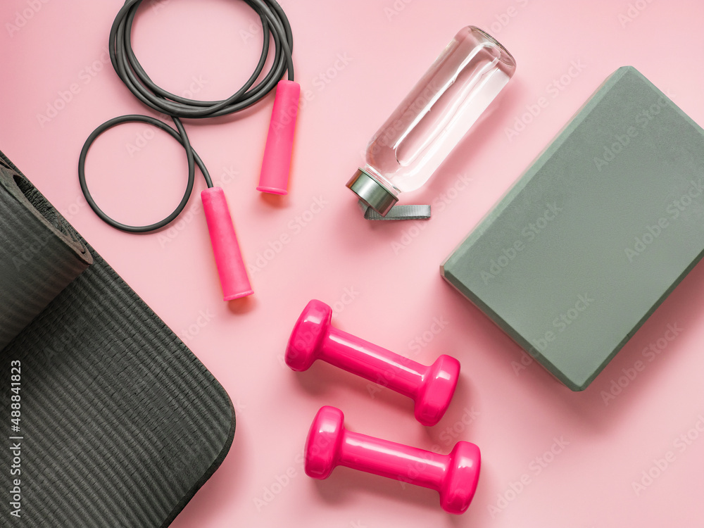 Naklejka premium Stylish gray and pink fitness training and gym flat lay. Top view of gray sport mat, yoga block, skipping rope, pink dumbbells, water bottle on pink background. Set for pilates, fitness, yoga