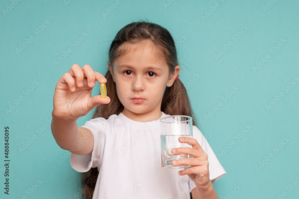 A sad little girl drinks a pill with water standing on a blue background A sad little girl drinks a pill with water standing on a blue background