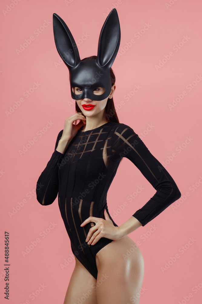 Sexy woman wearing a black mask Easter bunny standing on a pink background and looks very sensually