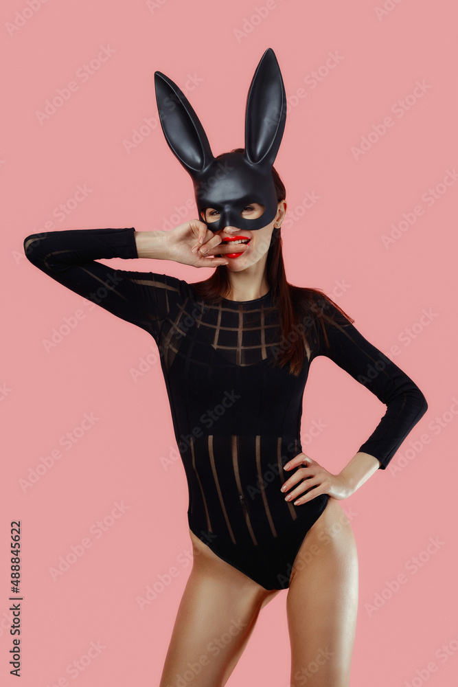 Sexy woman wearing a black mask Easter bunny standing on a pink background and looks very sensually