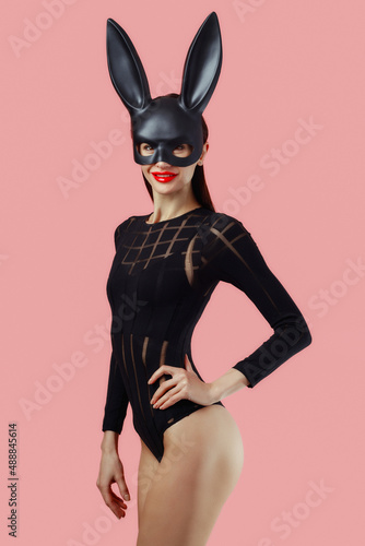 Sexy woman wearing a black mask Easter bunny standing on a pink background and looks very sensually