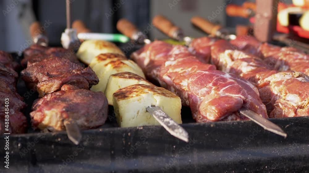 Juicy Appetizing Pork Shish Kebab, Feta Cheese Preparing in Smoke Outdoors. Cooking fresh meat on skewers, on hot coals in a grill in the sunlight at sunset. Trade, sale of fast food in the market. Juicy Appetizing Pork Shish Kebab, Feta Cheese Preparing in Smoke Outdoors. Cooking fresh meat on skewers, on hot coals in a grill in the sunlight at sunset. Trade, sale of fast food in the market.