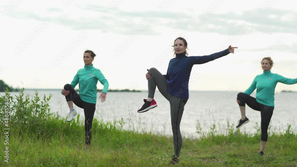 Tracking shot of group of mature women practicing yoga outdoors. Three women doing standing open knee pose in nature, yoga instructor in the middle Tracking shot of group of mature women practicing yoga outdoors. Three women doing standing open knee pose in nature, yoga instructor in the middle