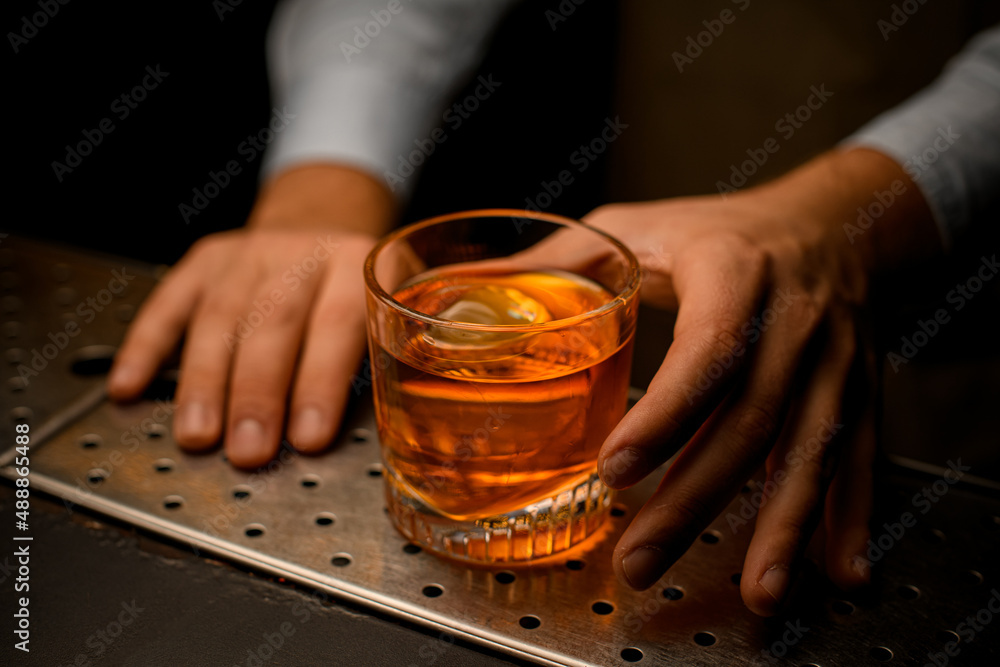 Cocktail glass with cold drink and ice cube stands on the bar counter and hand of male barman holds it