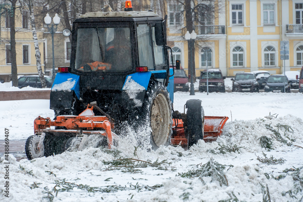 A tractor cleans snow in the city in winter after a snowfall. Cleaning city streets from snow. Tractor driver at work in the city square during a snowstorm.