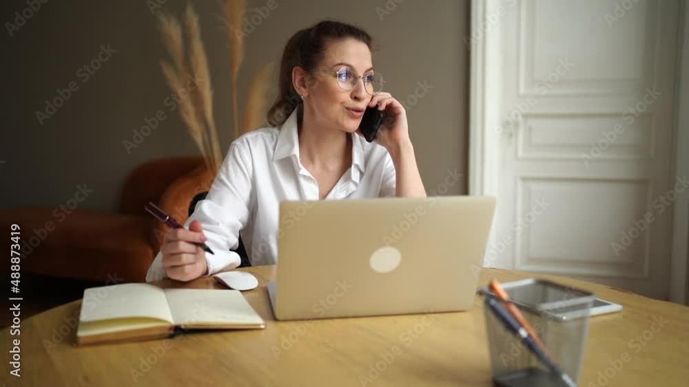 A young business woman with glasses in the office uses a phone call, makes a monthly report on a project on a laptop computer A young business woman with glasses in the office uses a phone call, makes a monthly report on a project on a laptop computer