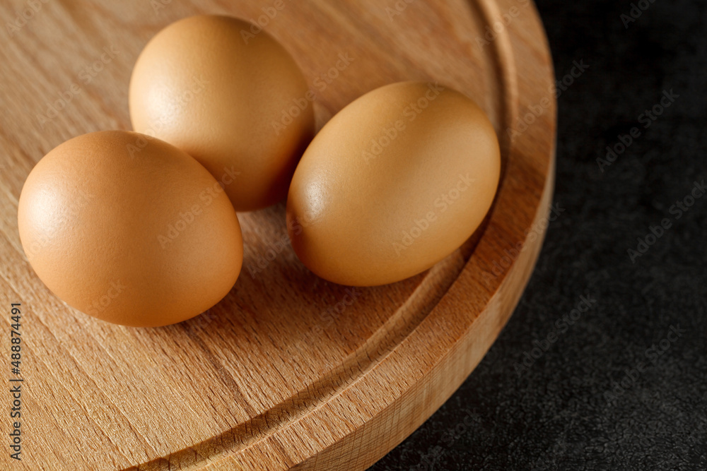 Eggs, three whole, lying on wooden board on dark background, selective focus. Eggs, three whole, lying on wooden board on dark background, selective focus.