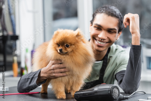 blurred african american man smiling near spitz and hair dryer on grooming table.