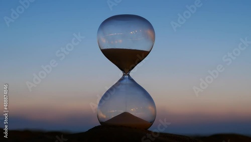 Hourglass time lapse shot, evening sky on background. Quickly night falls, picture becomes dark. Slowly at first, and then faster and quite quickly, remains of sand from upper bulb falls down