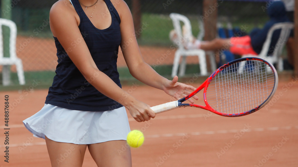 Jovem mulher rebatendo bola de tenis com camisa preta e saia azul e raquete vermelha