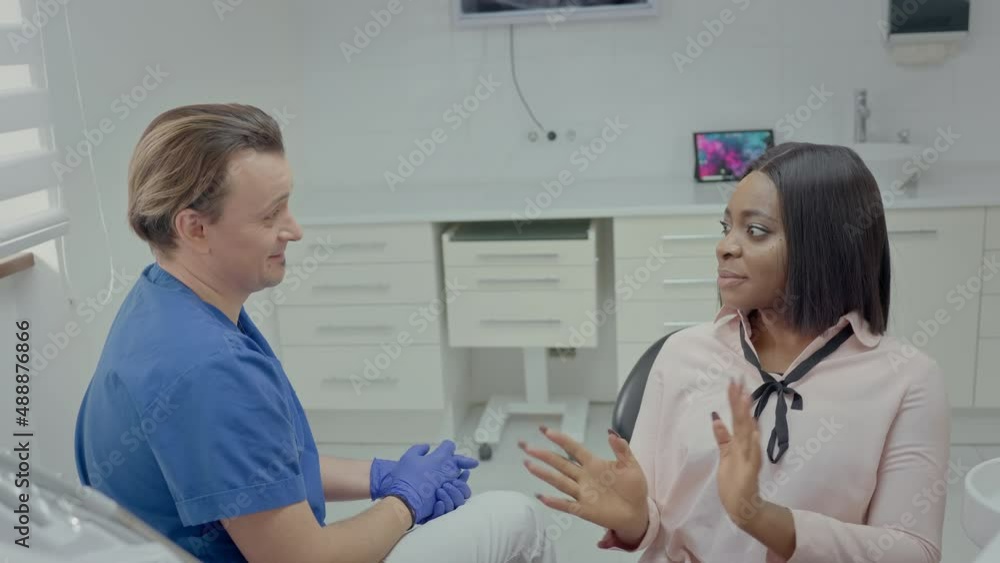 Reception At Dentist. Young Male Dentist Talking To Patient. In Chair Sits Black Young Girl. Doctor Laughs. Reception At Dentist. Young Male Dentist Talking To Patient. In Chair Sits Black Young Girl. Doctor Laughs.