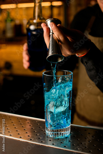 hand of the bartender pours blue beverage from jigger into mixing cup with ice