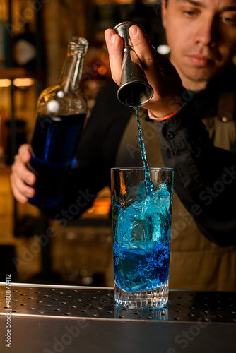 Close-up on glass with ice cubes into which bartender pours blue beverage from cocktail jigger.