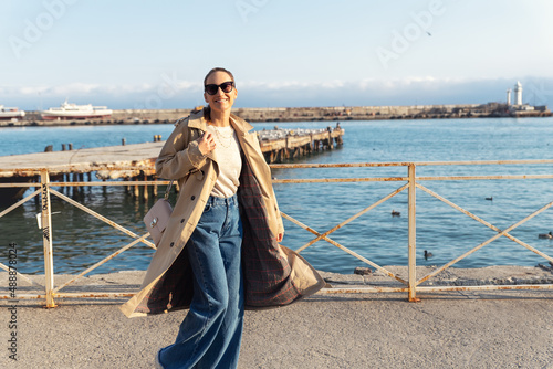 Young adult trendy stylish beautiful caucasian happy smiling woman enjoy walking by Yalta sea embankment on warm sunny day. Female person portrait wear jeans biege trench coat on urban city street