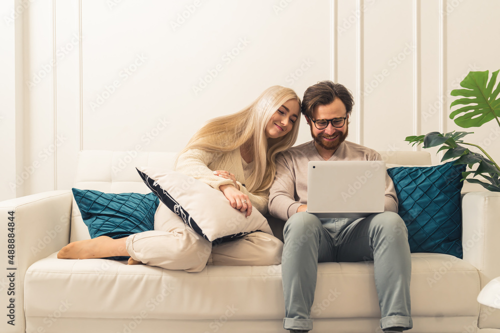 caucasian bearded man with laptop showing something funny at his blonde long-haired beautiful wife . High quality photo